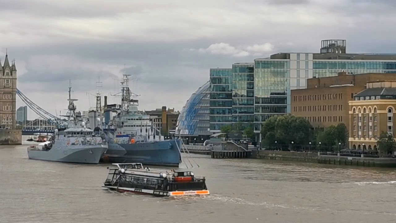 Grey warships moored by the south bank of the Thames as a passenger boat passes, with part of Tower Bridge on the left